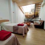 Habitación con dos camas individuales con sabanas blancas, marrones y rosas, un sofá, una mesa y un aire acondicionado.
